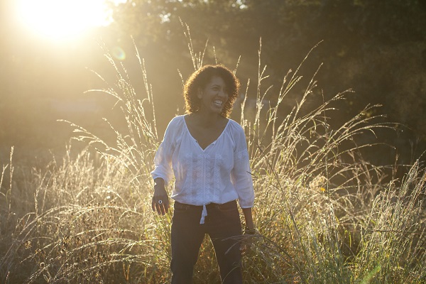 a woman is laughing as she stands in a field of tall grass. the sun rises behind her causing a halo effect on her and the grass.